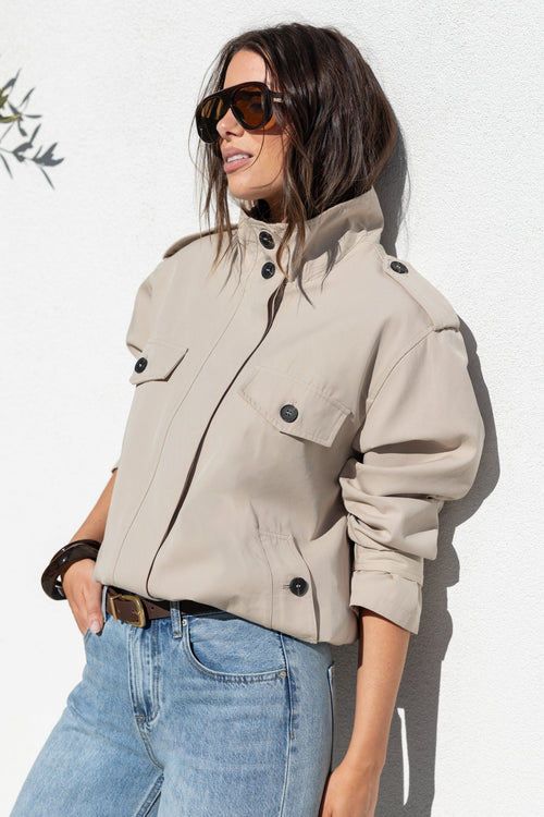 A woman wearing a light beige cropped utility jacket with black buttons, styled with blue jeans and sunglasses, posing against a white textured wall.