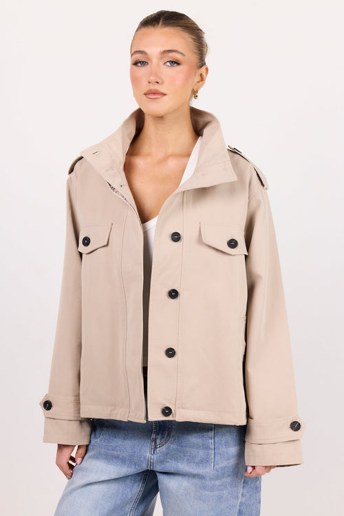 A woman modeling a light beige cropped military-style jacket with black buttons, worn over a white top and blue jeans, against a plain white studio background.