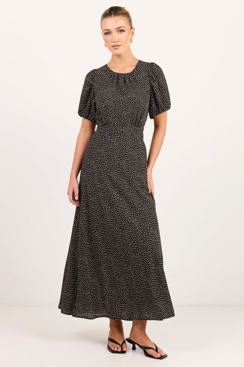 A woman modeling a short-sleeved black midi dress with a small white abstract pattern, standing against a plain white studio background.