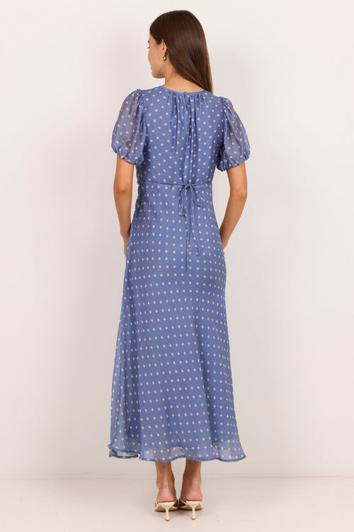A woman viewed from the back wearing a blue midi dress with a white polka dot pattern, short puff sleeves, and a gathered waist, standing against a plain off-white studio background.