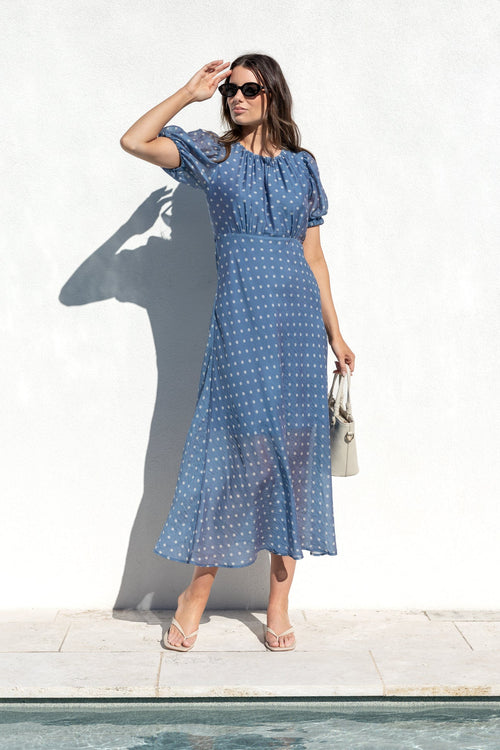 A woman wearing a blue and white patterned midi dress stands next to a swimming pool in front of a white wall, holding a small beige handbag.