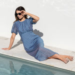 A woman wearing a blue short-sleeve midi dress with white polka dots and sunglasses reclines on the edge of a swimming pool against a white wall.