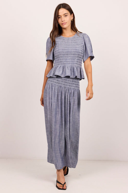 A woman modeling a matching two-piece set consisting of a smocked short-sleeve top with a ruffle hem and matching wide-leg pants in a textured slate blue fabric, set against a plain light background.