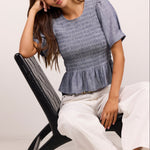 A woman models a blue-gray smocked short-sleeve peplum top paired with white cropped pants and black sandals, seated on a black modern chair against a plain off-white studio background.