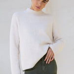 Woman wearing a white sweater and green pants standing on a sidewalk.