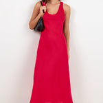 A woman modeling a sleeveless bright red midi dress with a scoop neckline, paired with black sunglasses, a black shoulder bag, and black flip-flop sandals, standing against a plain white studio background.