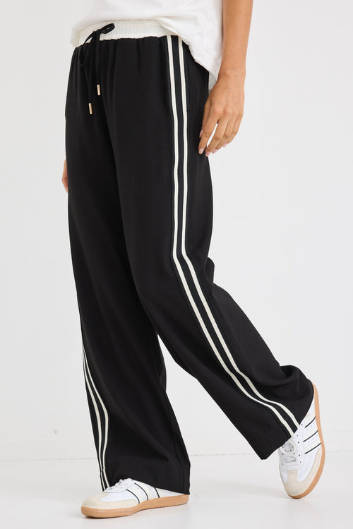 Woman wearing black wide-leg pants with white side stripes, featuring a mid-rise elastic waistband and paired with sneakers.