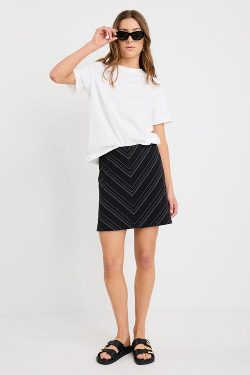 Woman wearing a black and white gradient stripe mini skirt in a chevron pattern, styled with a simple white tee and black sandals.