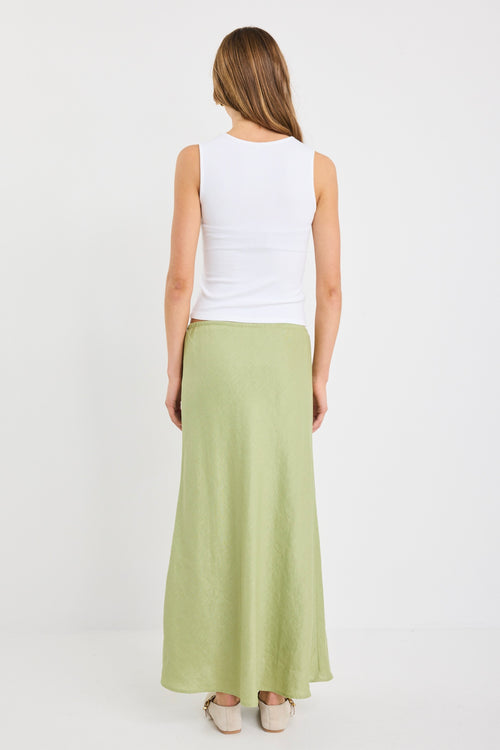 Woman wearing a plain moss green bias-cut maxi skirt with elastic waist and tie detail, styled with a white tank top and neutral flats.