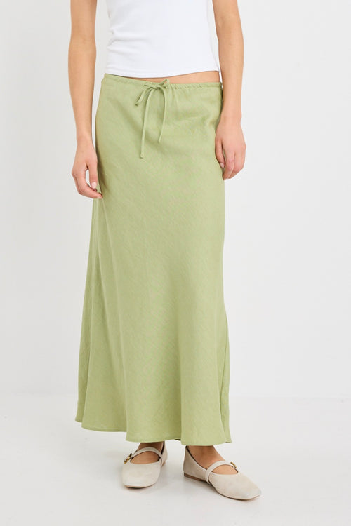 Woman wearing a plain moss green bias-cut maxi skirt with elastic waist and tie detail, styled with a white tank top and neutral flats.