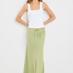 Woman wearing a plain moss green bias-cut maxi skirt with elastic waist and tie detail, styled with a white tank top and neutral flats.