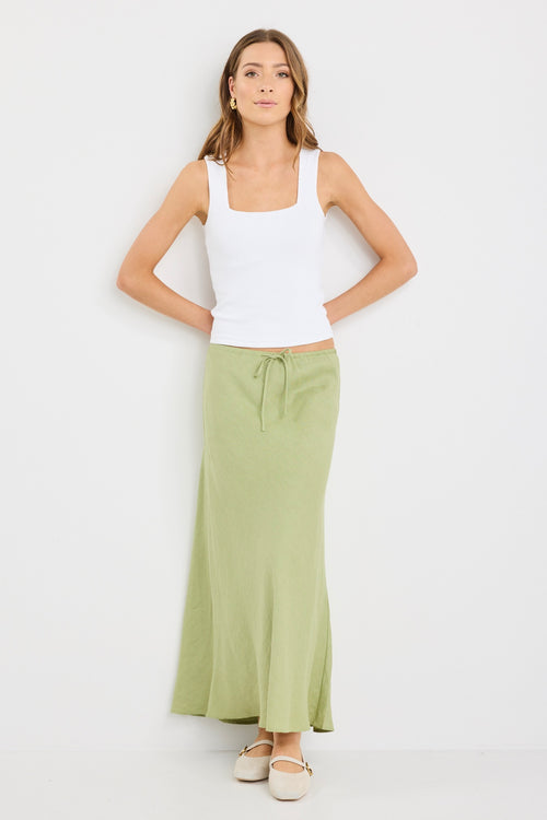 Woman wearing a plain moss green bias-cut maxi skirt with elastic waist and tie detail, styled with a white tank top and neutral flats.