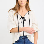 Woman wearing a cream long sleeve linen top with black trim and tie-front detail, designed in an oversized fit with puff sleeves, paired with blue jeans.