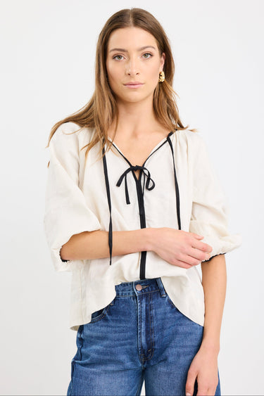Woman wearing a cream long sleeve linen top with black trim and tie-front detail, designed in an oversized fit with puff sleeves, paired with blue jeans.