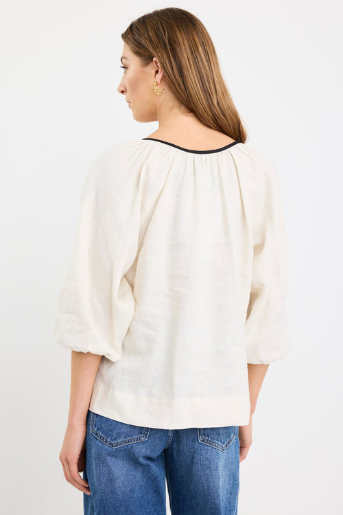 Woman wearing a cream long sleeve linen top with black trim and tie-front detail, designed in an oversized fit with puff sleeves, paired with blue jeans.