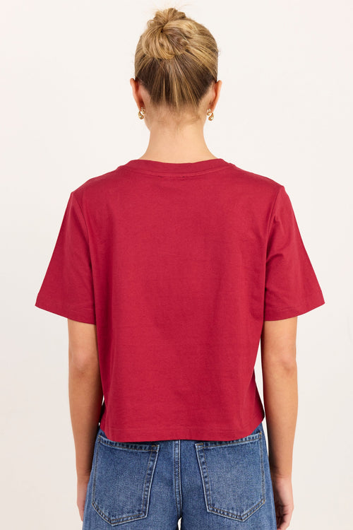 A woman viewed from behind wearing a short-sleeve cropped t-shirt in a deep red color, paired with blue jeans, against a plain off-white background.