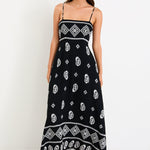 Woman wearing a black maxi dress with fine straps, white paisley and border print, and shirred back detail.