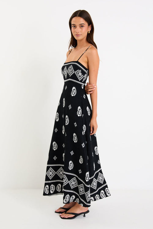 Woman wearing a black maxi dress with fine straps, white paisley and border print, and shirred back detail.