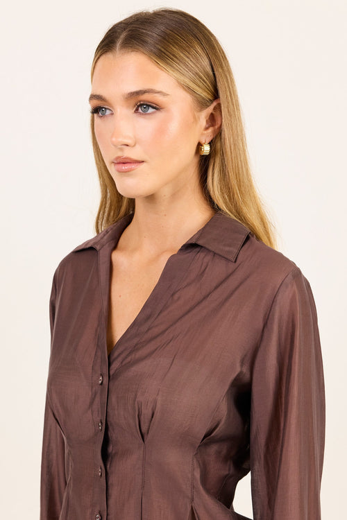 A woman modeling a brown long-sleeve button-up shirt with a V-neck and gathered side details, photographed against a plain light background.