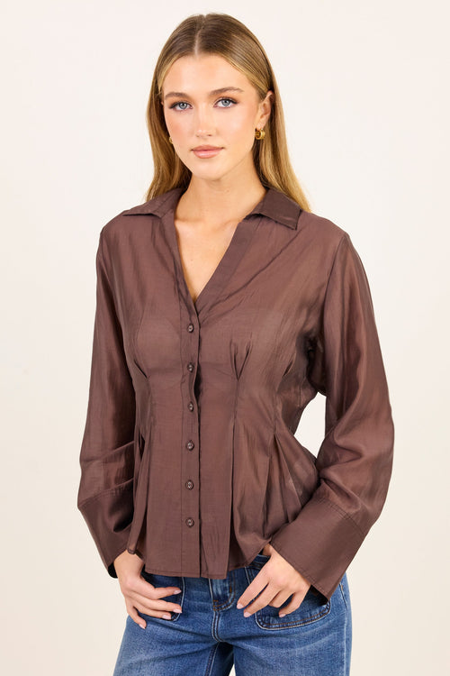 A woman modeling a brown long-sleeve button-up blouse with a V-neck and gathered front, paired with blue jeans, against a plain off-white studio background.
