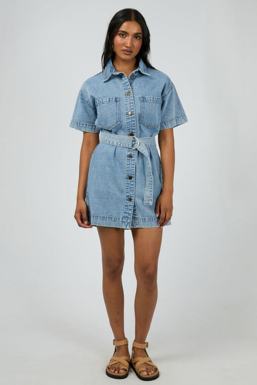 Woman wears a short-sleeve light wash denim mini dress with front buttons, patch pockets, and a matching fabric belt cinched at the waist for a structured, feminine silhouette.