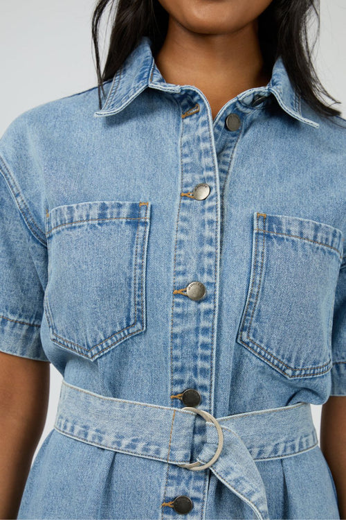 Woman wears a short-sleeve light wash denim mini dress with front buttons, patch pockets, and a matching fabric belt cinched at the waist for a structured, feminine silhouette.