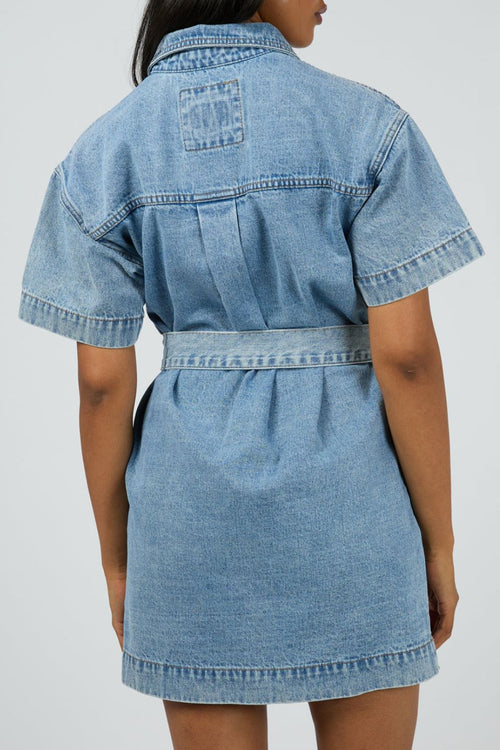 Woman wears a short-sleeve light wash denim mini dress with front buttons, patch pockets, and a matching fabric belt cinched at the waist for a structured, feminine silhouette.