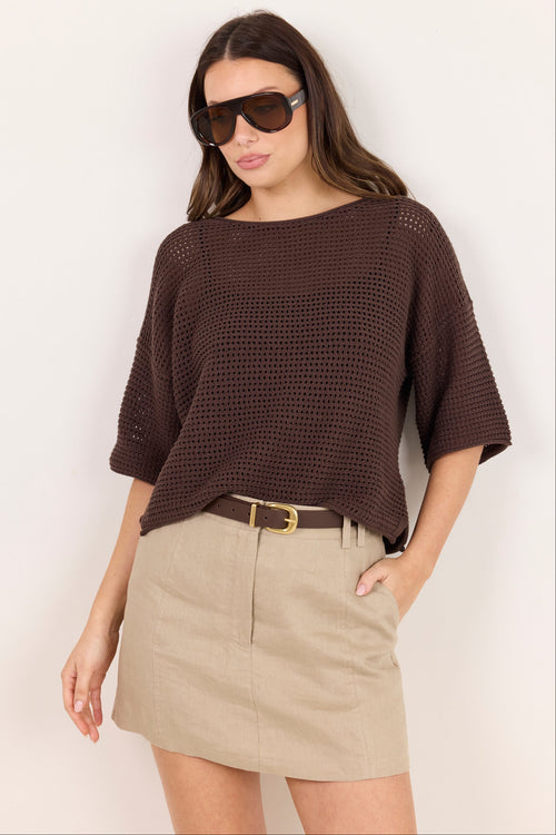 A woman modeling a dark brown open-knit short-sleeve top with a boat neckline, paired with a beige mini skirt and sunglasses, against a plain off-white background.