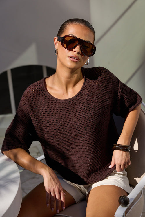 A woman wearing a dark brown open-knit short-sleeve top and beige shorts, sitting on a white chair in a modern setting with strong sunlight and shadows.