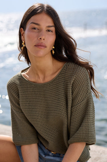 A woman sits on a concrete pier by the water wearing a loose olive green knit top, denim shorts, and black flip-flops.