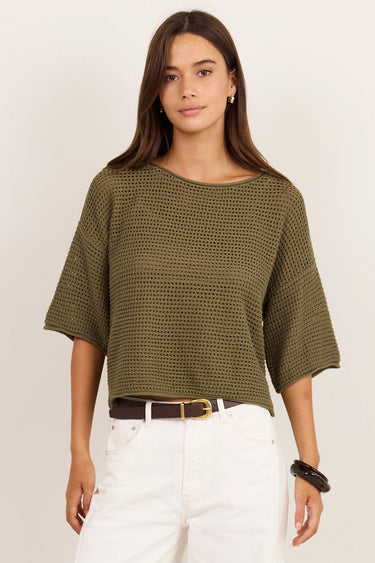 A woman modeling a short-sleeve olive green ribbed knit top with white wide-leg pants, a brown belt, and black sandals, standing against a plain off-white studio background.