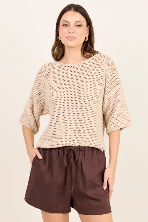 A woman modeling a beige open-knit cropped sweater with elbow-length sleeves and dark brown drawstring shorts against a plain cream background.