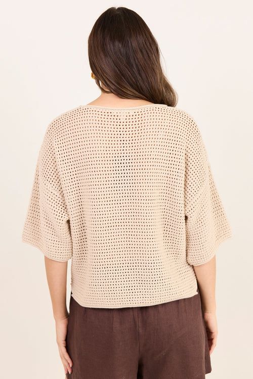 A woman viewed from the back wearing a beige open-knit cropped top with elbow-length sleeves and dark brown pants, against a plain off-white background.
col_pattern