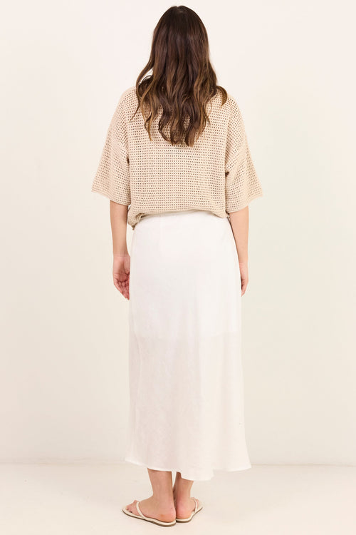 A woman viewed from behind wearing a beige textured short-sleeve top and a long white skirt, standing against a plain off-white studio background.