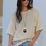 A woman wearing a cream-colored open-knit short-sleeve top, light-wash blue jeans, and black sunglasses, standing against a white stucco wall.