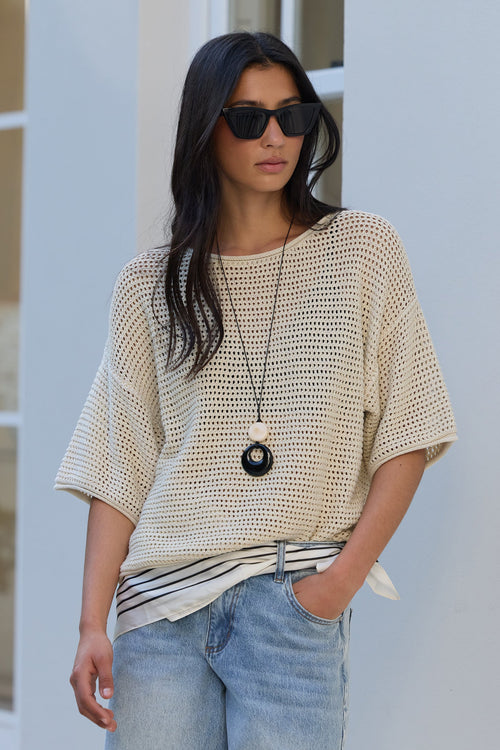 A woman wearing a cream-colored open-knit short-sleeve top, light-wash blue jeans, and black sunglasses, standing against a white stucco wall.