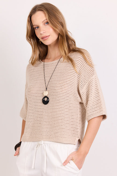A woman wearing a beige open-knit short-sleeve top and white drawstring pants, accessorized with a long black pendant necklace, against a plain light background.