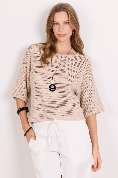 A woman modeling a beige open-knit short-sleeve top with white drawstring pants, standing against a plain white background.