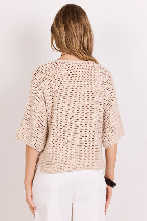 A woman viewed from behind wearing a beige open-knit cropped sweater with elbow-length sleeves and white pants, standing against a plain light background.