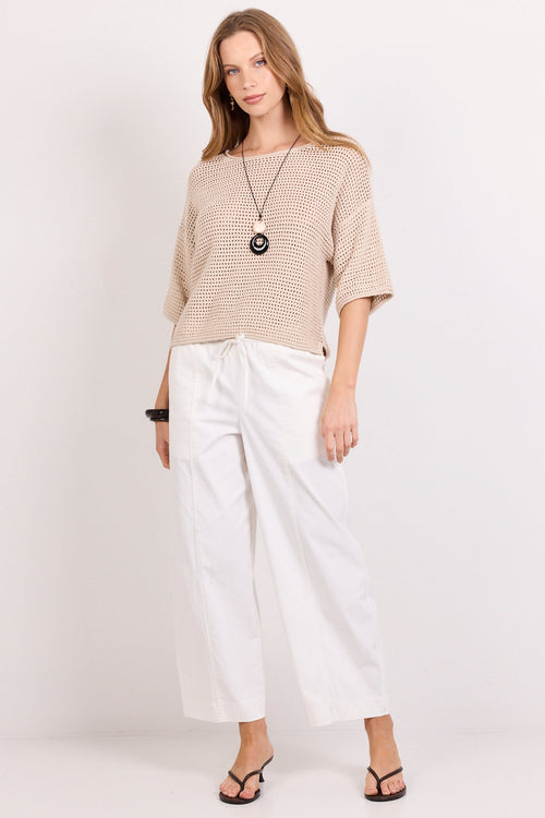A woman modeling a beige knit short-sleeve top and white wide-leg cropped pants against a plain white studio background.