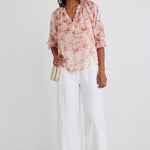 Model wears a floral blouse with white linen pants and brown sunglasses. 