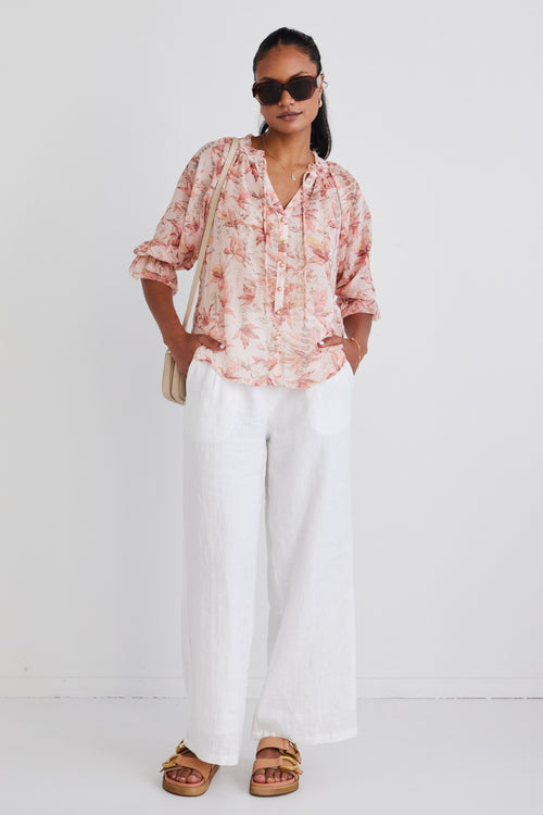 Model wears a floral blouse with white linen pants and brown sunglasses. 