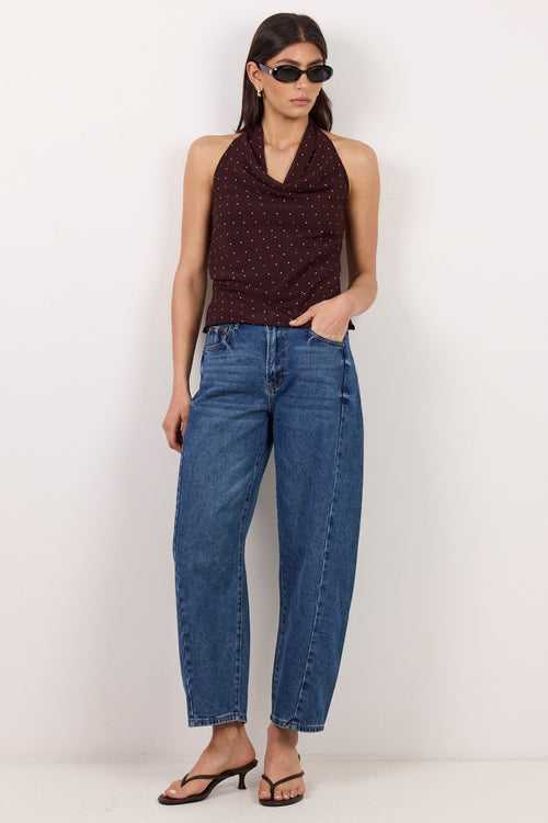 A model stands against a plain white studio wall wearing a dark brown halter top with a small red pattern, blue wide-leg cropped jeans, and black sandals.
