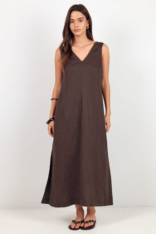 A woman modeling a sleeveless, dark brown linen maxi dress with a V-neckline, standing against a plain white studio background.