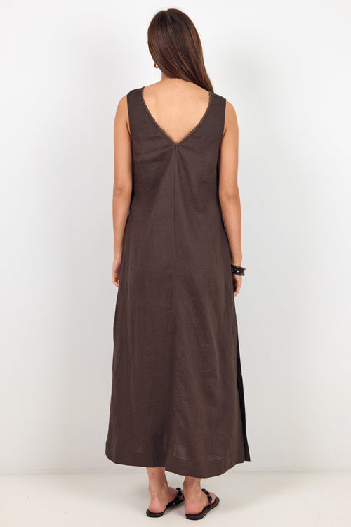 A woman viewed from behind wearing a sleeveless, dark brown maxi dress with a deep V-neckline and side slits, standing against a plain white studio background.