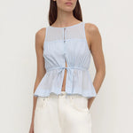 A woman modeling a light blue sleeveless top with a sheer yoke, button front, and gathered peplum hem, paired with white pants, against a plain light gray studio background.