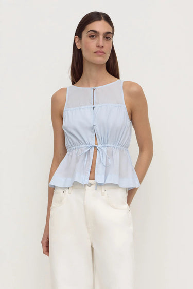 A woman modeling a light blue sleeveless top with a sheer yoke, button front, and gathered peplum hem, paired with white pants, against a plain light gray studio background.