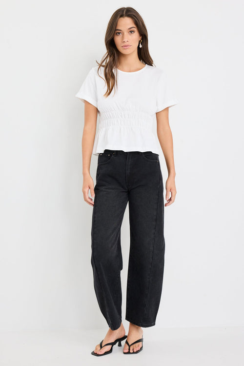 Woman wearing an ivory short sleeve cotton tee with shirred waist detail and a subtle peplum hem, styled with black jeans.