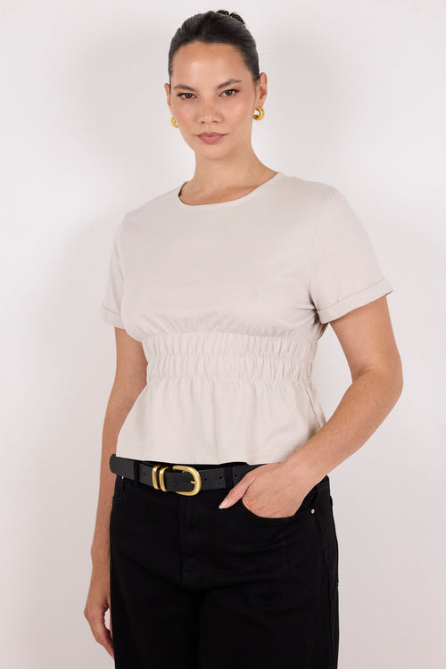A woman modeling a short-sleeve crewneck top in a light beige or off-white color, featuring a gathered elastic waist. She is wearing black trousers with a black belt and gold buckle, set against a plain light gray studio background.