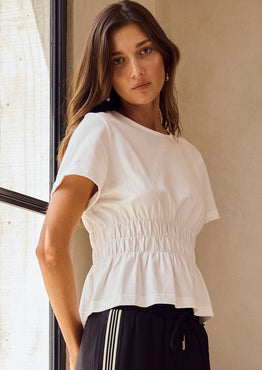 Woman in a white top and black pants with white stripes standing by a large window.